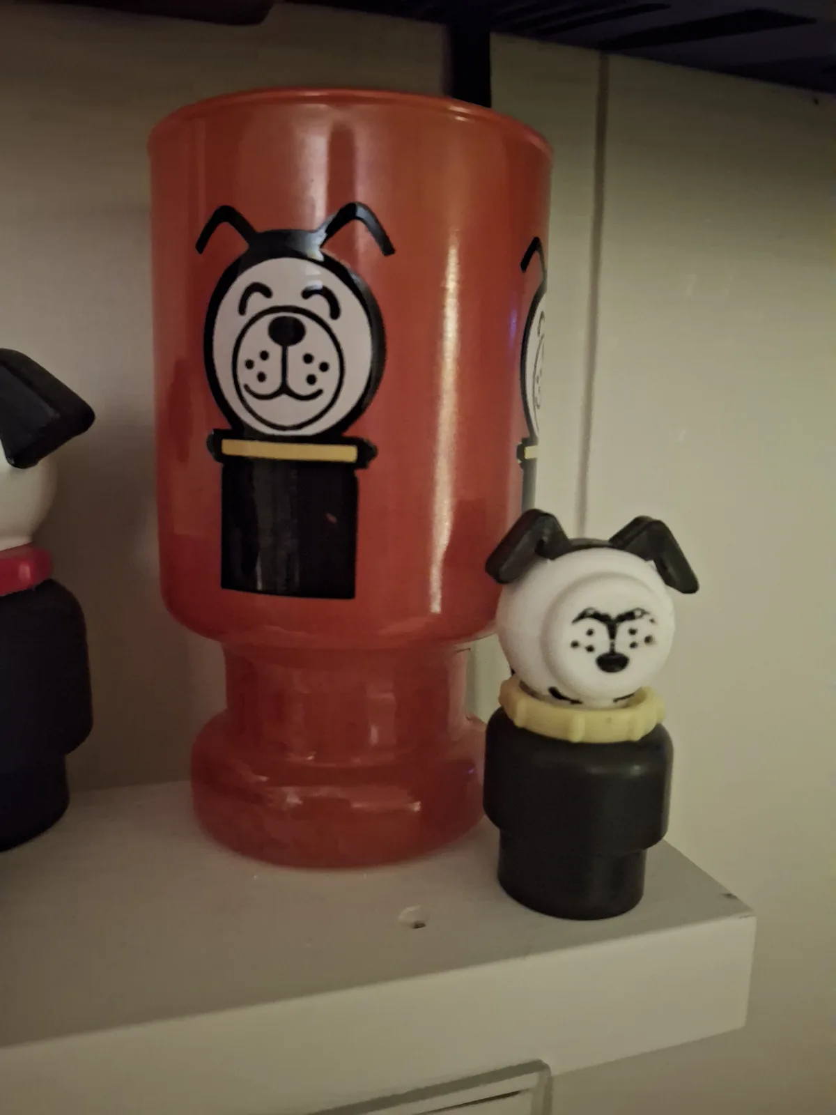 A small dog figurine with an angry face stands next to a large orange dog-themed cup with a smiling dog face, both on a…