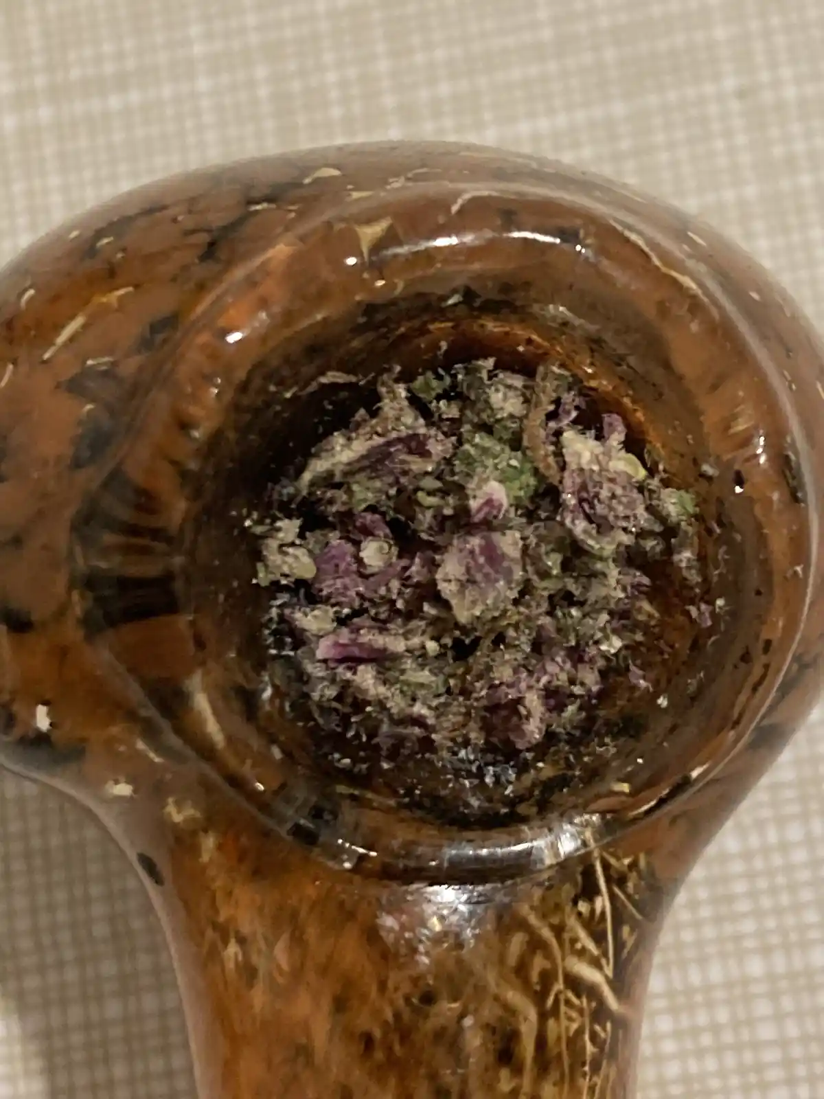 A close-up photo shows the top of a brown glass pipe filled with ground, dried plant material, likely cannabis, with visible…