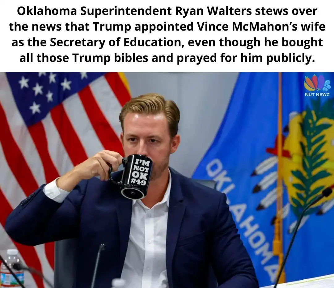 A man in a dark suit and white shirt sits at a table with microphones, drinking from a black mug that reads 'I'M NOT OK…