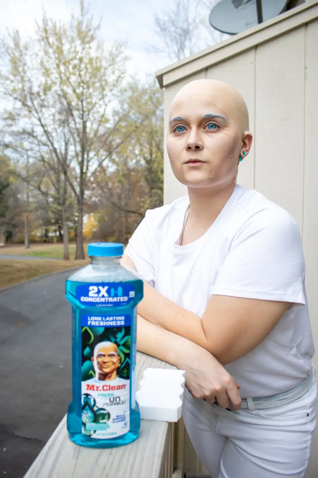 A person with a bald head, blue eyes, and white clothing stands beside a bottle of Mr. Clean cleaner, mimicking the iconic…