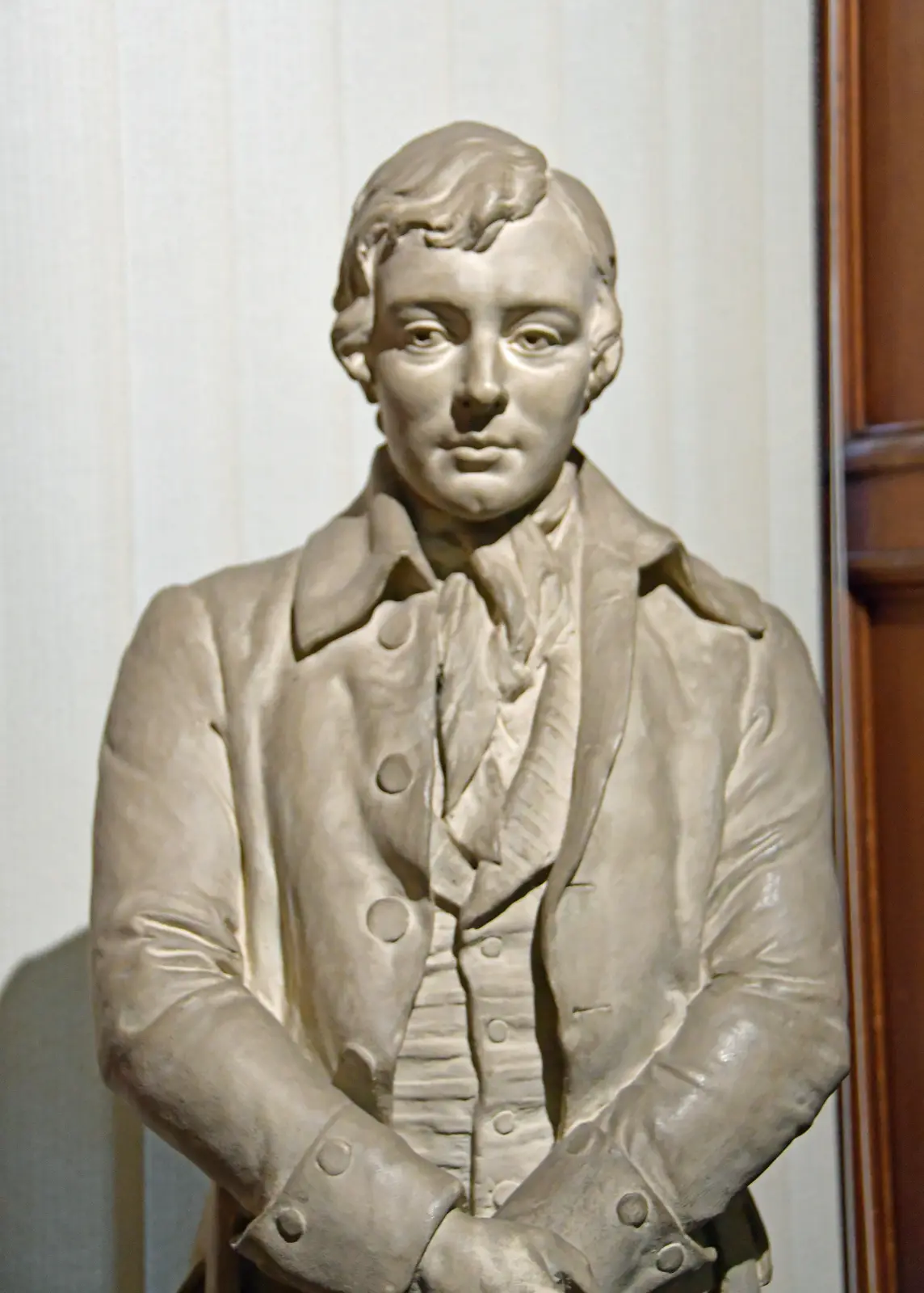 A marble bust of a man in 18th-century formal attire, including a coat with buttons and a cravat, stands against a plain…
