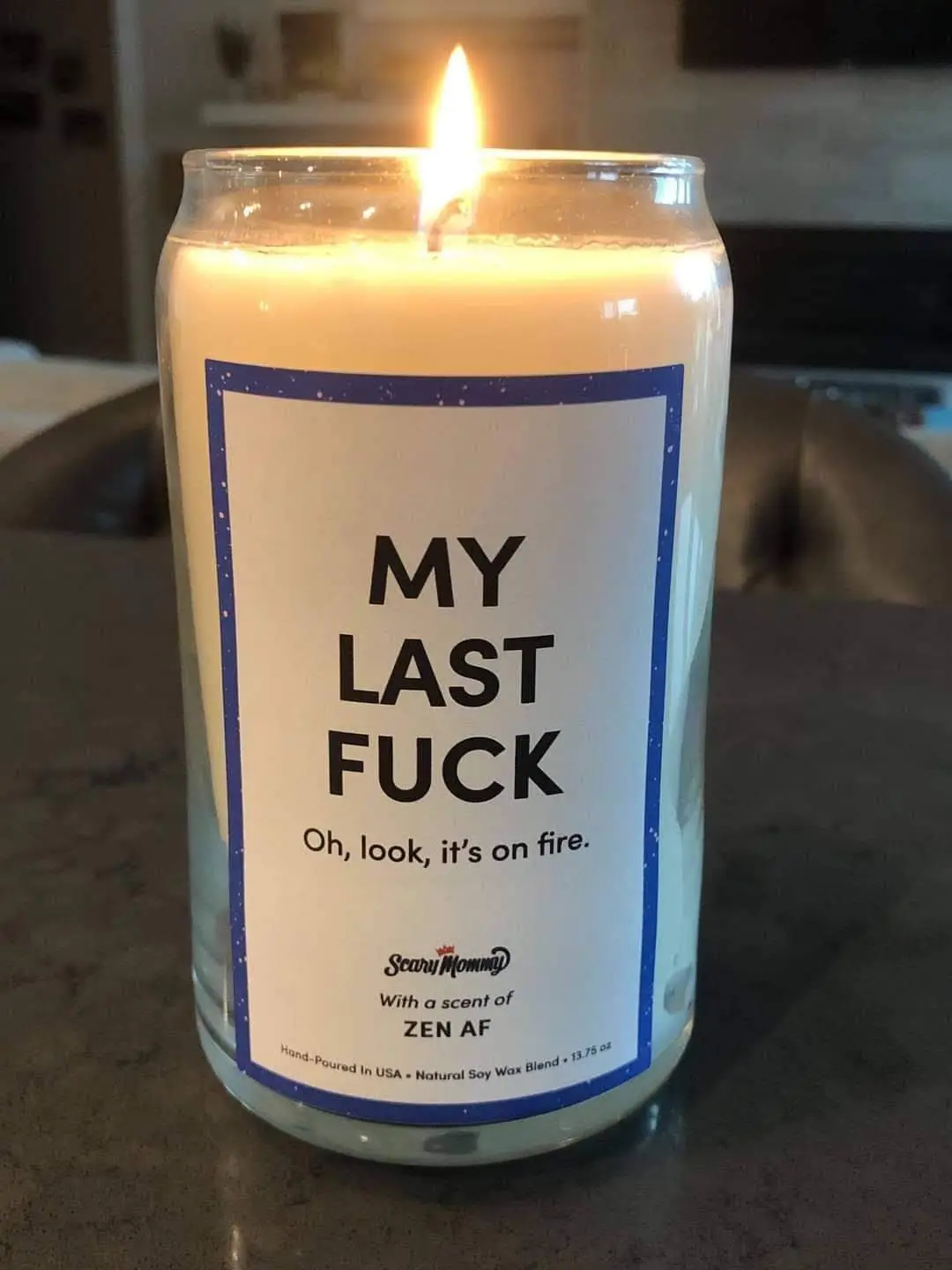 A lit candle in a clear glass jar with a white label that reads 'MY LAST FUCK' and 'Oh, look, it's on fire.' The label also…