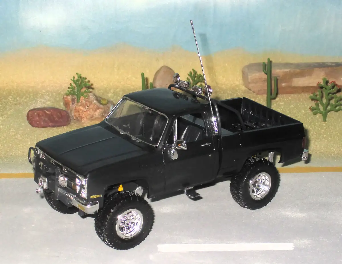 A black toy pickup truck with oversized tires, chrome rims, roof lights, and a tall antenna is parked on a gray surface with…