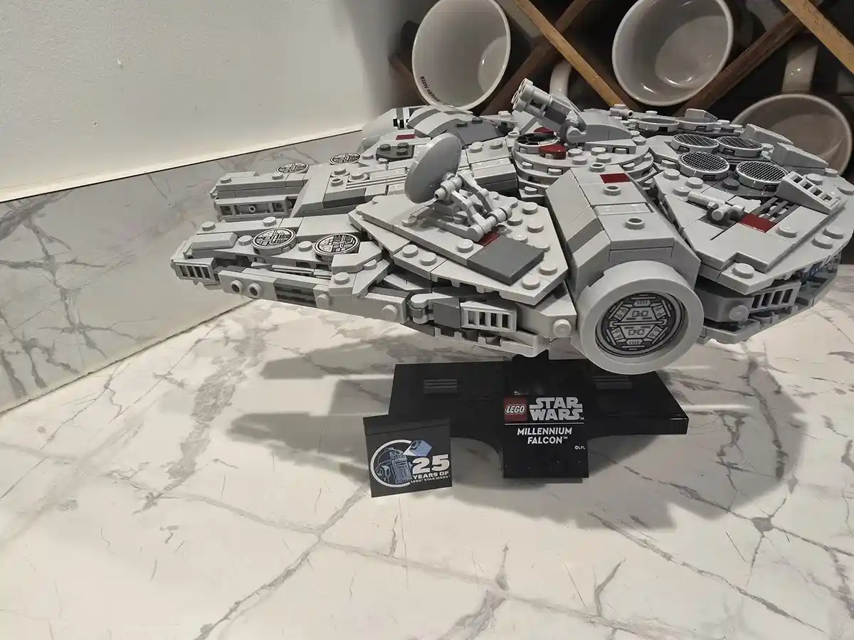 A large, detailed LEGO Millennium Falcon model sits on a black display stand on a white marble countertop, with a mug rack…