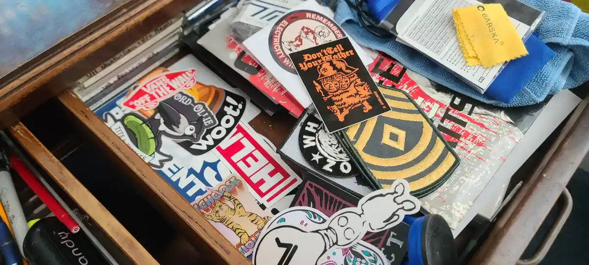 A cluttered wooden drawer filled with various stickers, patches, and small items, including a prominent black sticker with…