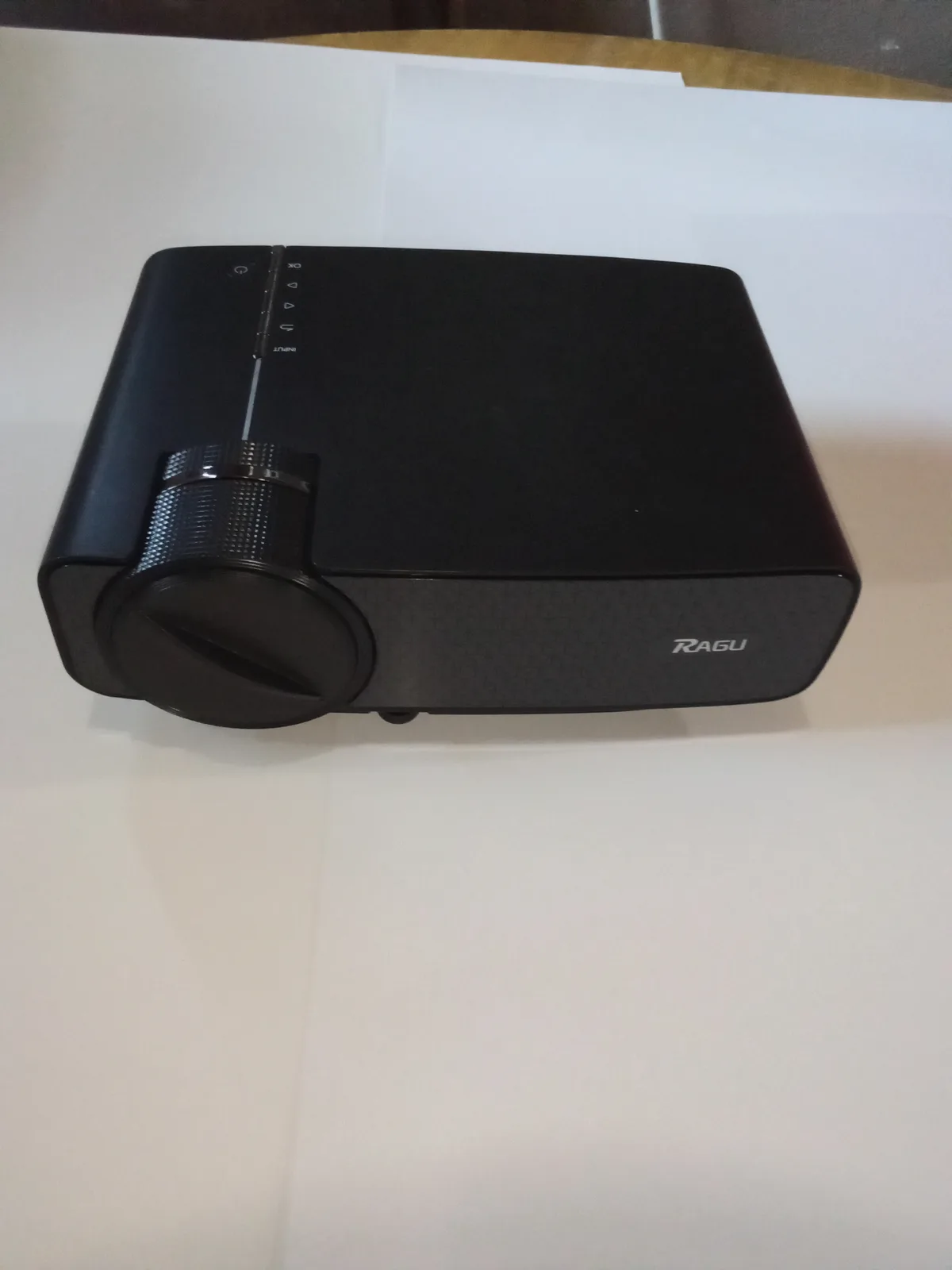 A black RAGU brand projector sits on a white surface, showing its lens and control buttons. The overlaid text reads 'i…