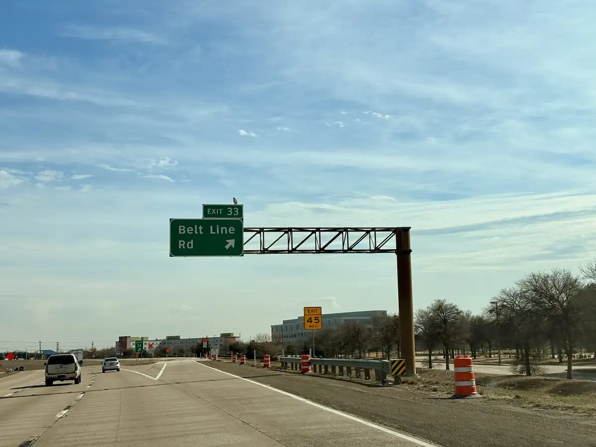 A bird is perched atop a green highway sign indicating Exit 33 for Belt Line Rd, with a yellow 45 MPH speed limit sign…