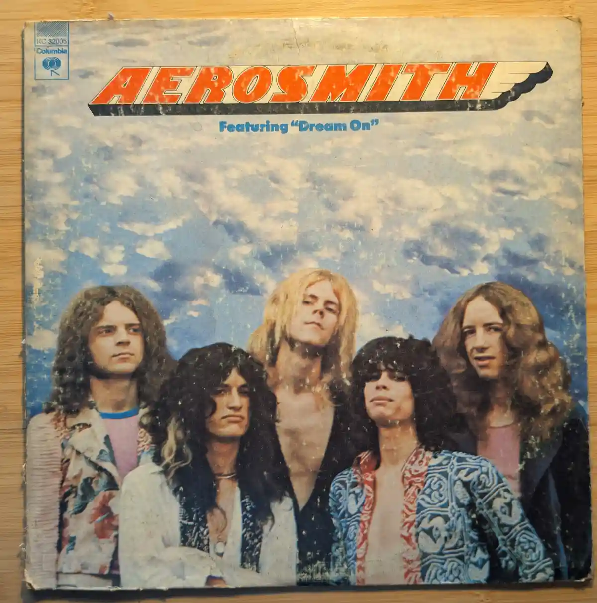 A weathered, vintage Aerosmith record sleeve featuring the band's classic 1970s photo against a cloudy sky, with the band…