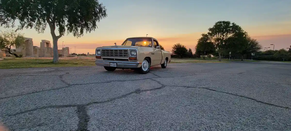 A beige 1970s Dodge pickup truck is parked in an empty asphalt lot at sunrise, with the sun visible through the windshield…