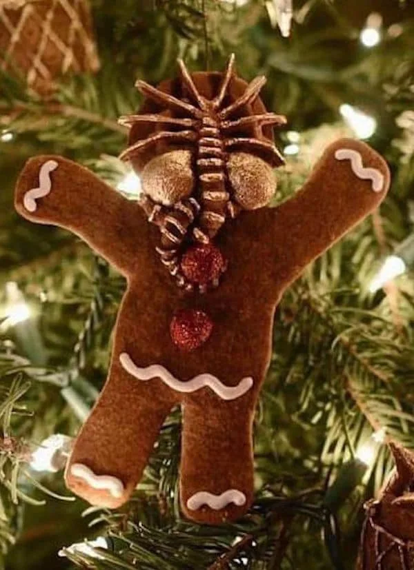 a brown felt gingerbread man ornament hangs on a lit christmas tree, its head replaced with a pinecone and its mouth a red…