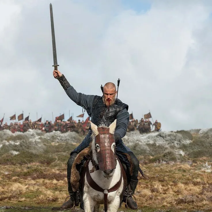 A bald, bearded man in medieval armor rides a white horse with a leather muzzle, holding a sword high in the air. He has…
