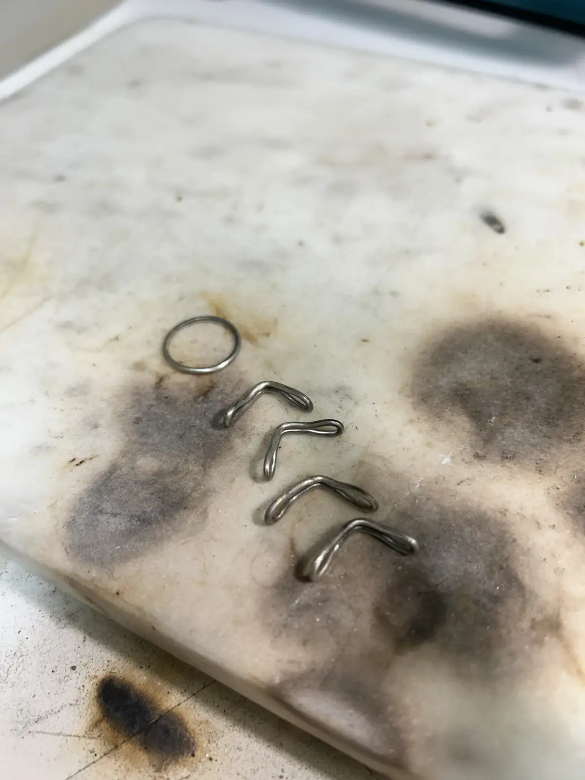A close-up photo shows five small, bent metal pieces arranged vertically on a dirty, stained white marble surface. The top…