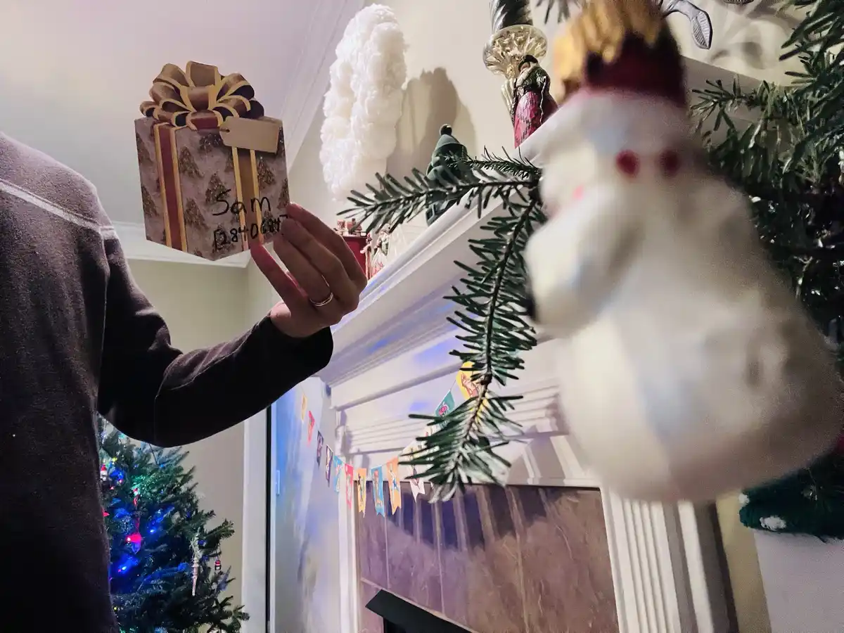 A person holds a small, decorated gift box labeled 'Sam 12840687' in front of a fireplace decorated with Christmas…