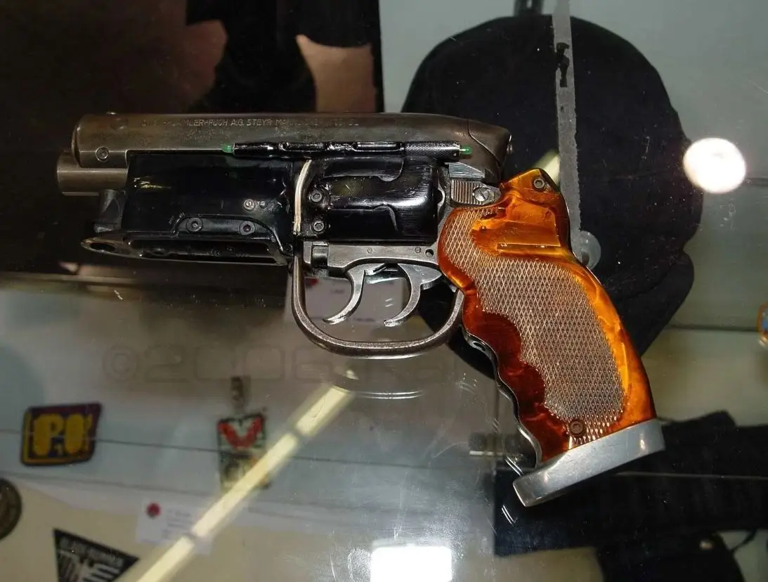 a steyr m1912 pistol with a distinctive amber-colored grip is displayed inside a glass case, with a black cap visible behind…