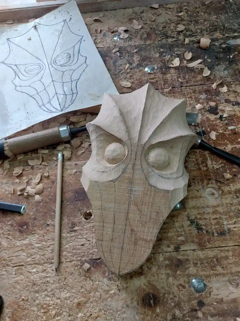 A partially carved wooden mask sits on a workbench surrounded by wood shavings, carving tools, and a pencil. A paper sketch…