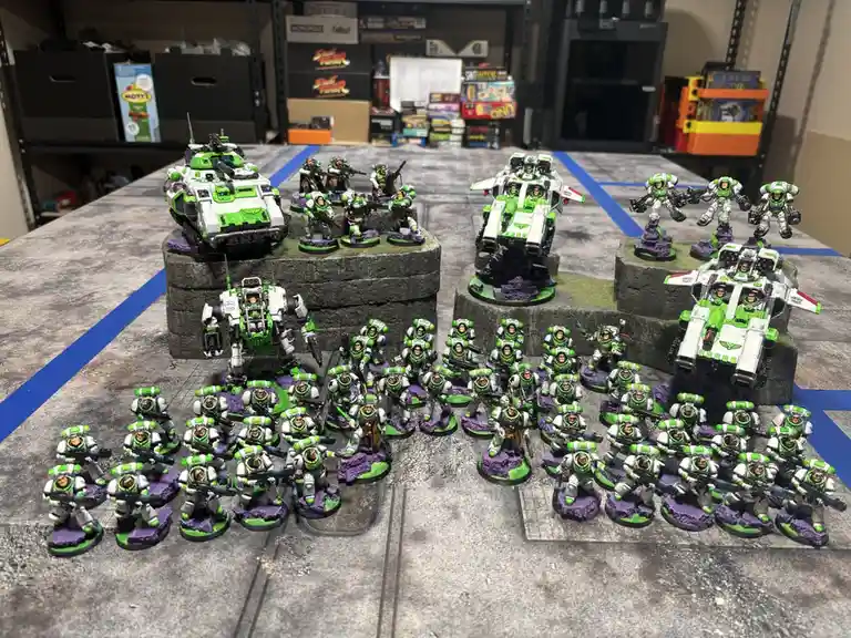 A large collection of painted miniature figurines, mostly in green and white armor, arranged on a gray tabletop with blue…