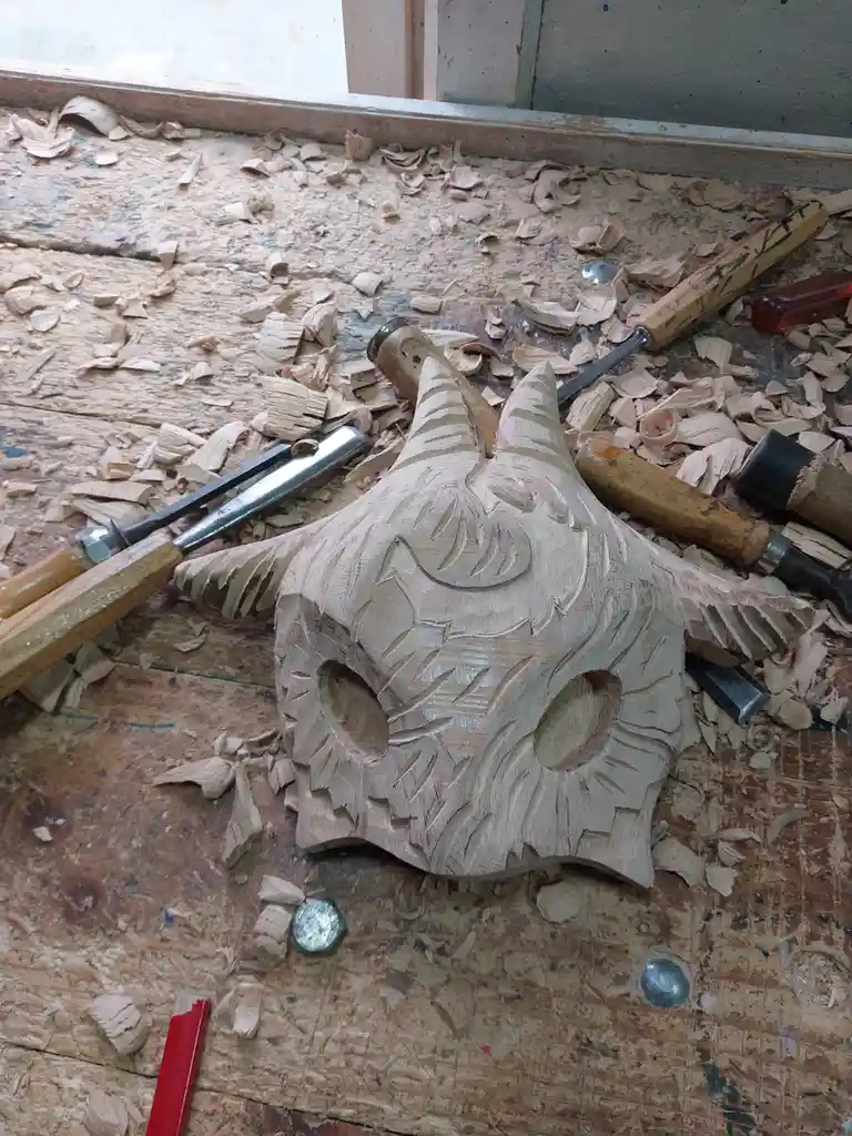 A partially carved wooden mask with horns and facial features sits on a workbench covered in wood shavings, surrounded by…