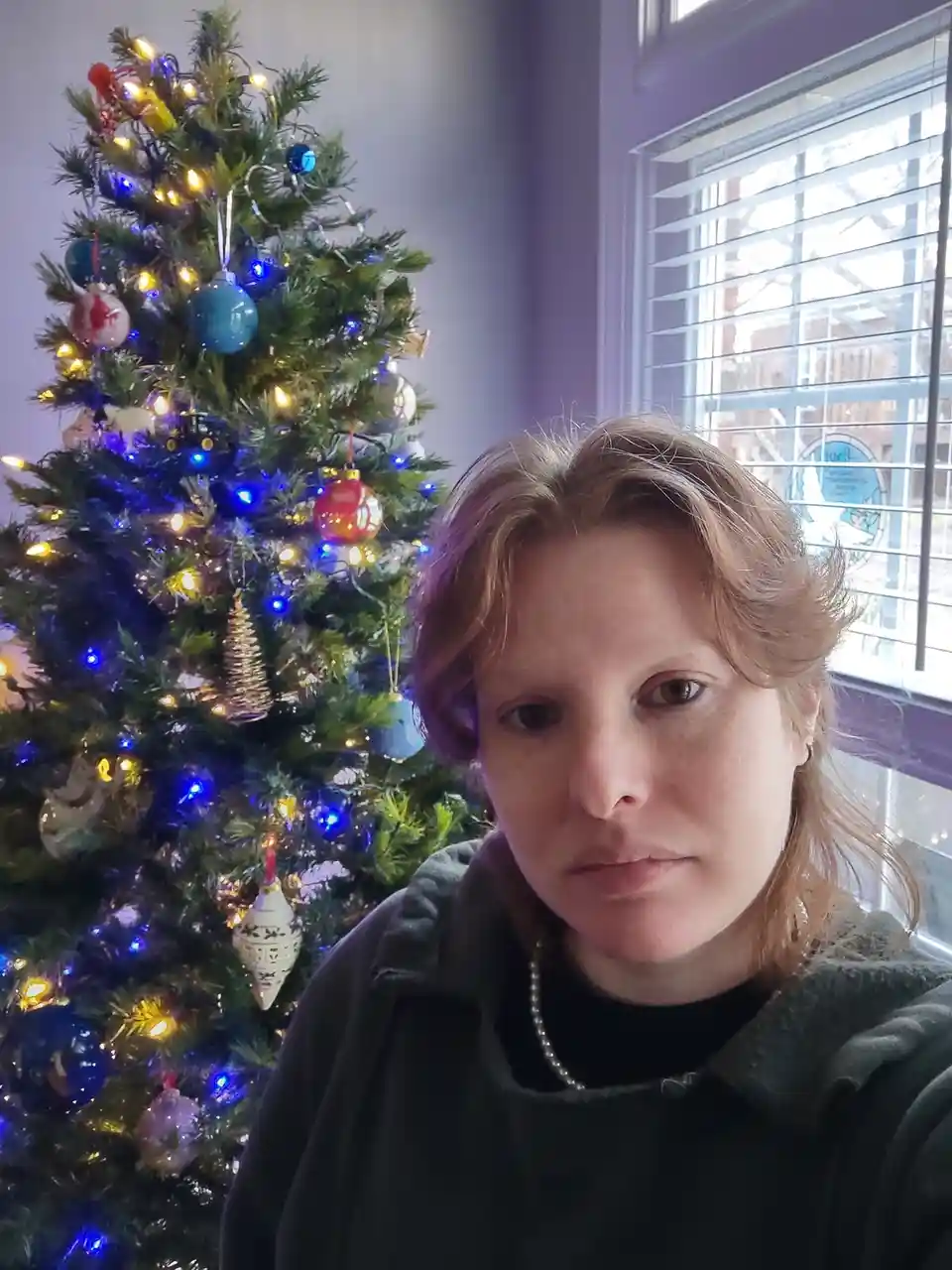A woman with reddish-brown hair stares directly at the camera with a deadpan, unamused expression. Behind her is a brightly…