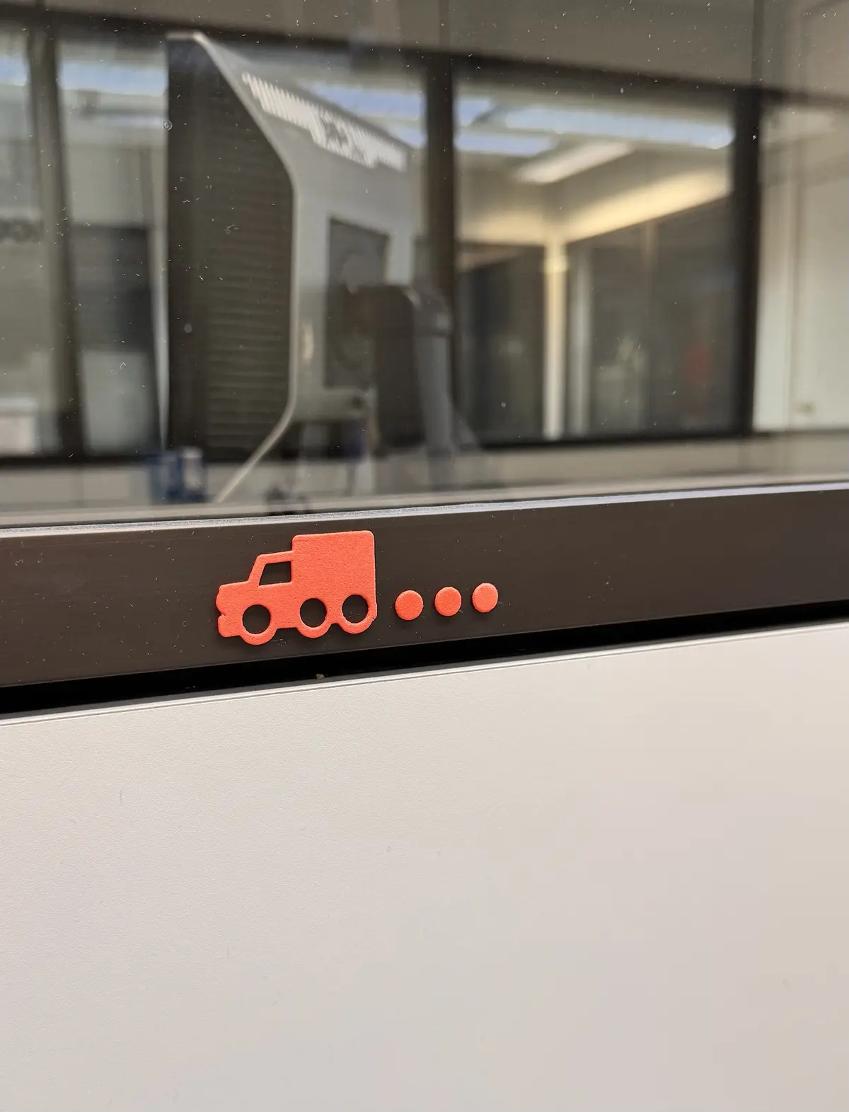 A close-up photo shows a small, orange, 3D-printed truck icon sticker affixed to a dark brown horizontal strip on a white…