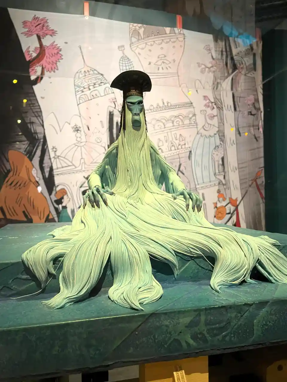 A green, long-haired, humanoid statue with a monkey-like face and a black hat sits regally on a green platform. Behind it is…