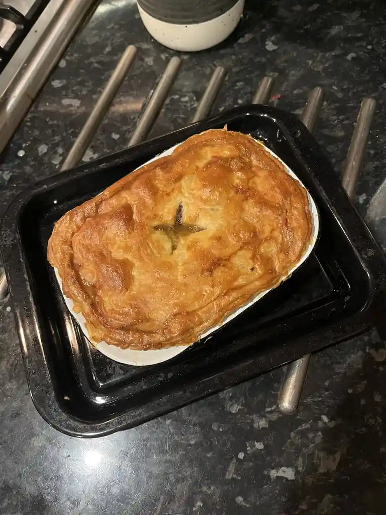 A golden-brown, freshly baked pie sits in a white dish on a black baking tray, placed on a dark kitchen countertop. The…