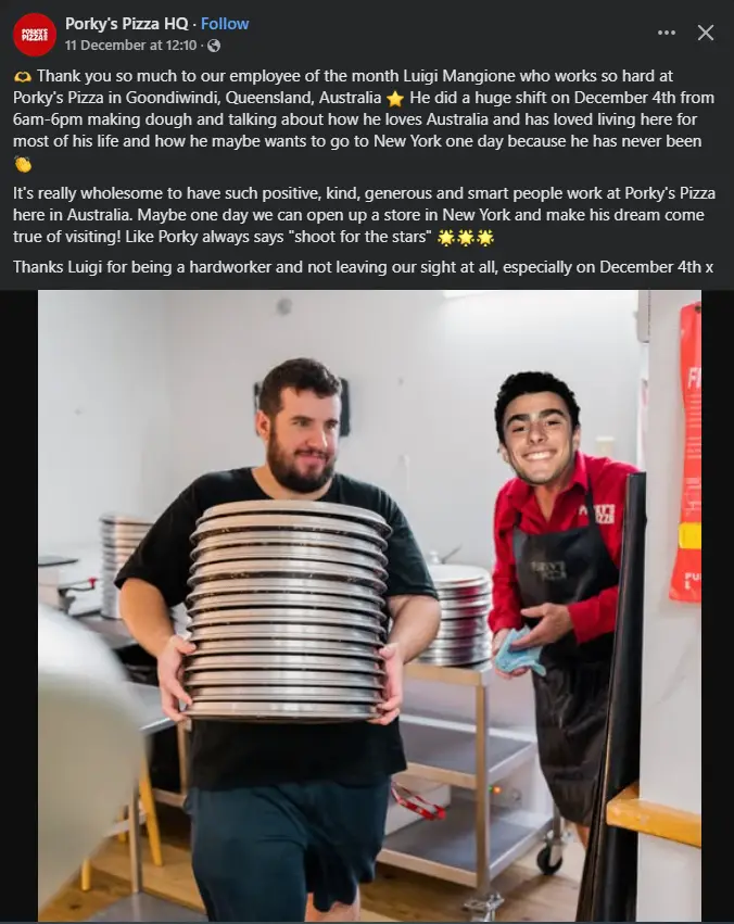 Two men in a pizza kitchen, one carrying a tall stack of metal pizza pans, the other smiling beside him. The image is…