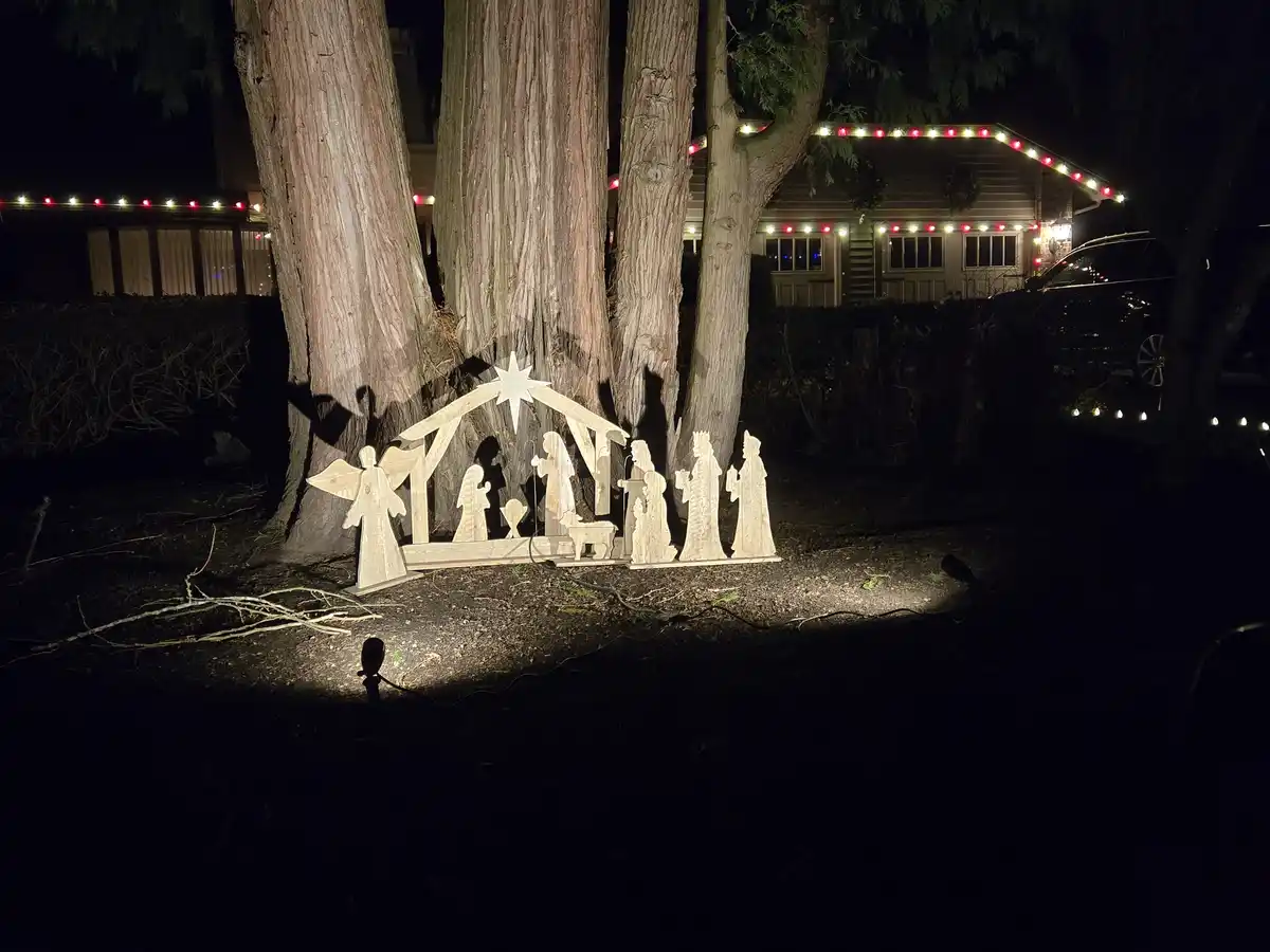 A nighttime photo shows a wooden nativity scene set up outdoors under large trees, illuminated by a spotlight. The scene…