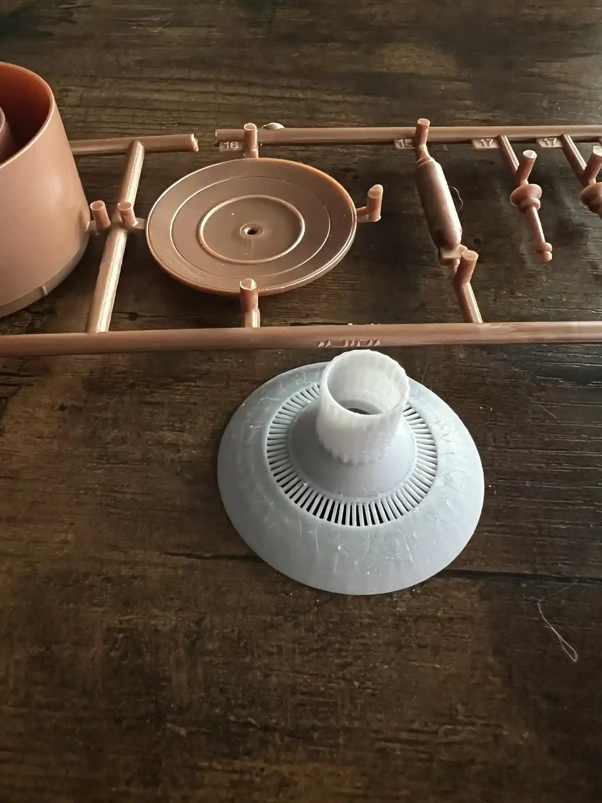 A close-up photo shows a 3d-printed white plastic part with a fluted base and central tube, sitting on a dark wooden…