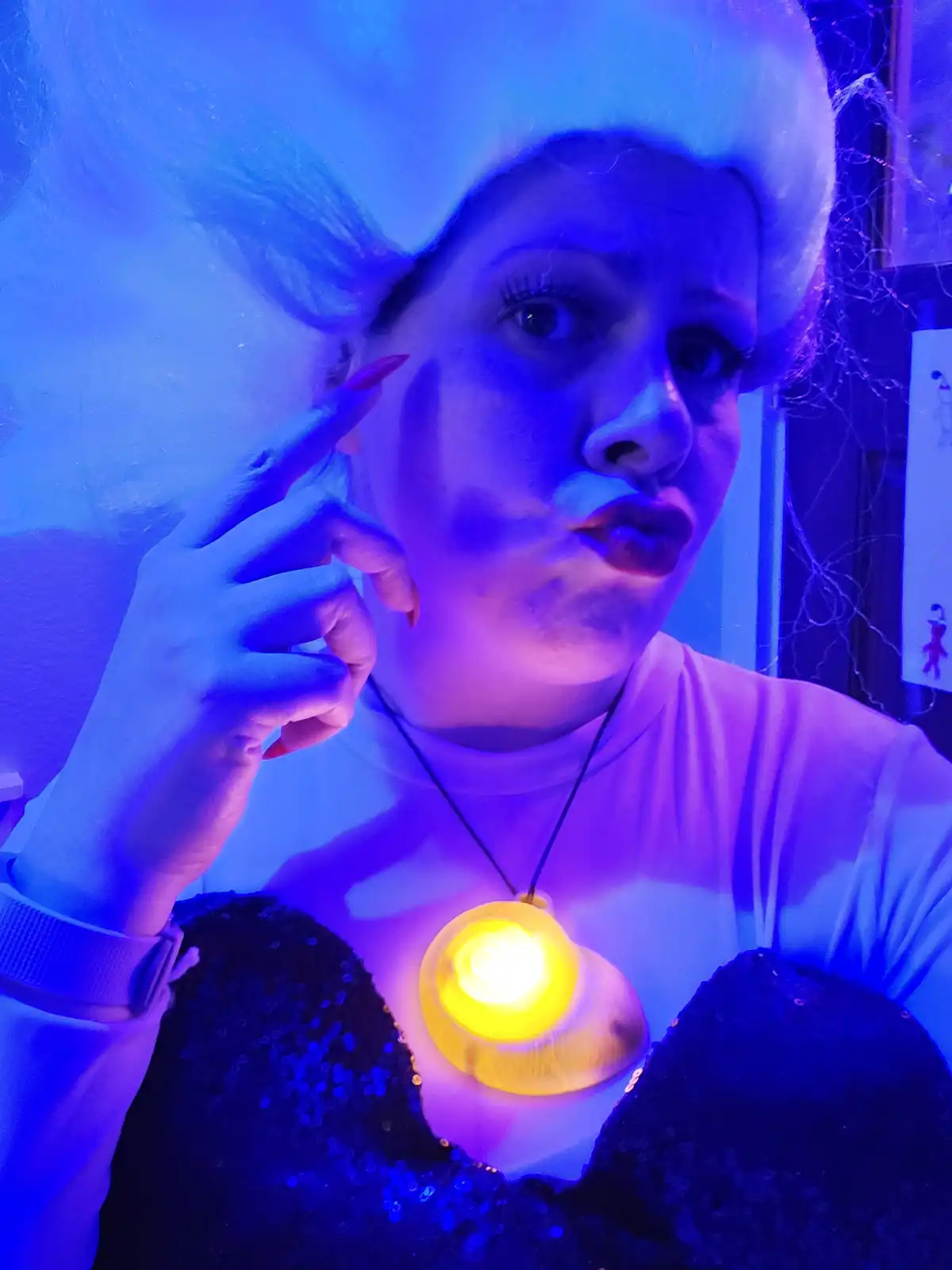 A person in a mermaid costume with a blue wig and sequined top holds a glowing yellow lightbulb pendant necklace while…