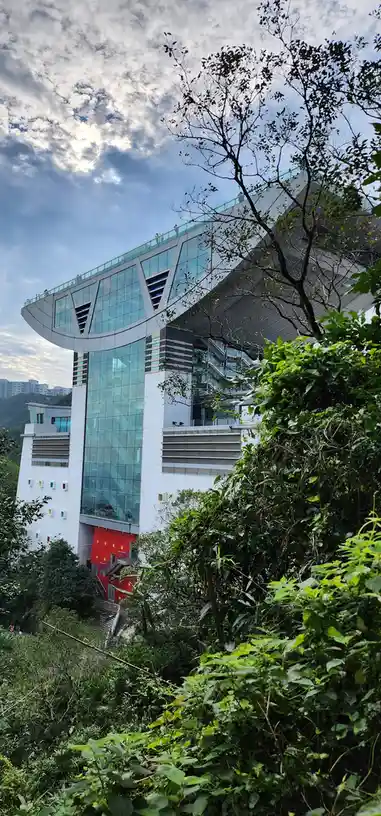 A modern glass and white building with a curved rooftop terrace, partially obscured by lush green foliage in the foreground…