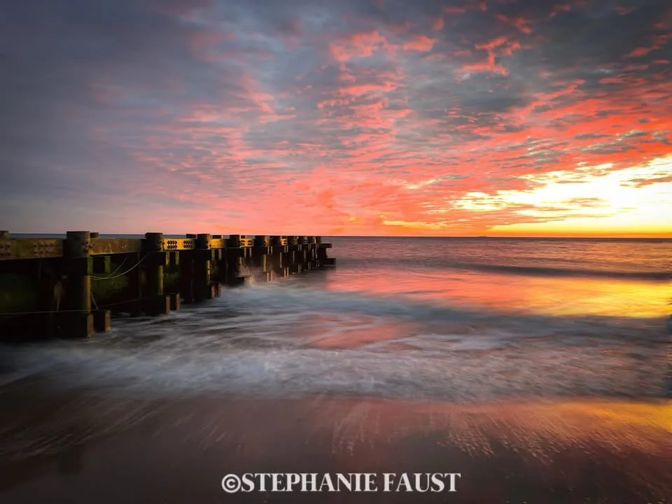 A long-exposure photograph of a serene beach at sunset, featuring a wooden pier extending into the ocean under a sky painted…
