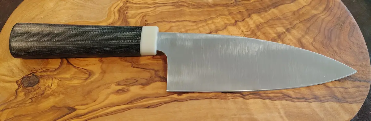 A chef's knife with a dark textured handle and a white bolster rests on a wooden cutting board with prominent grain…