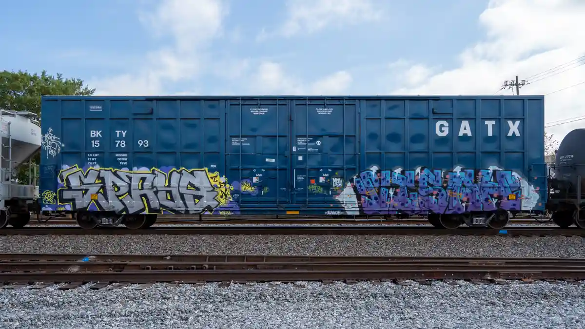A blue GATX freight train car sits on tracks under a partly cloudy sky, covered in colorful graffiti tags. The overlaid text…