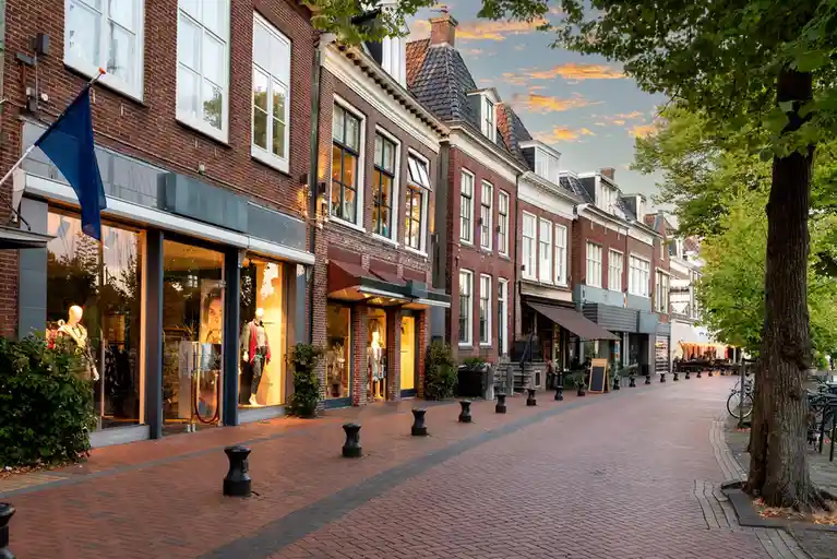 A picturesque European street lined with brick buildings, shops with mannequins in windows, and outdoor café seating under…