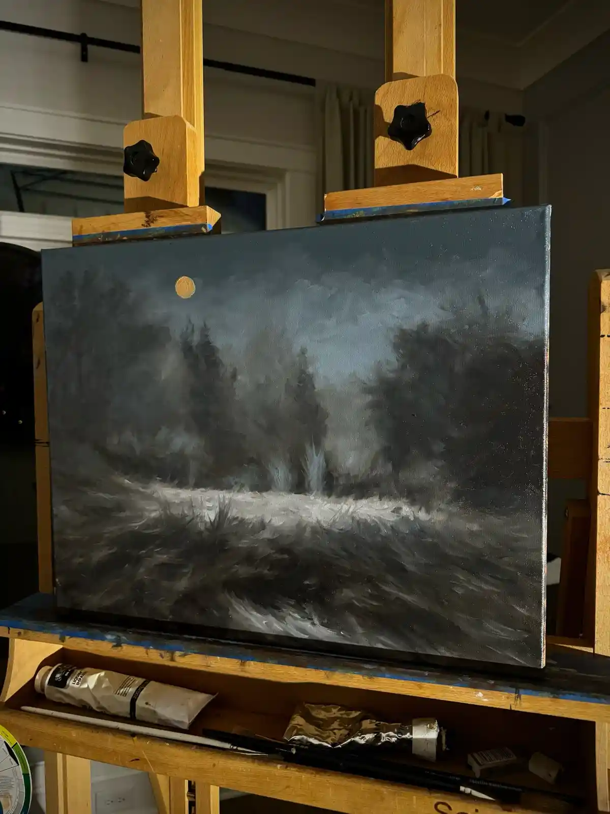 A painting on a wooden easel depicts a dark, moody forest scene with a single, small, golden moon in the sky. The canvas is…
