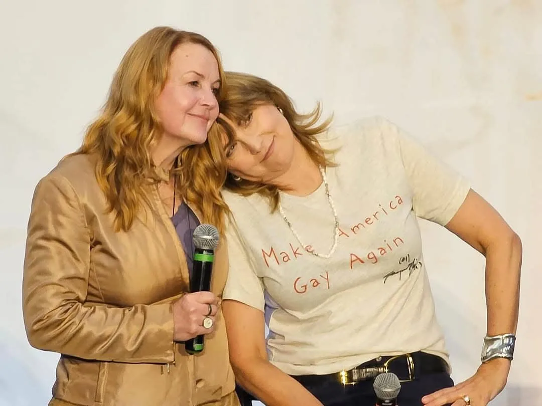 Two women are posing together on a stage; one holds a microphone and wears a tan jacket, while the other leans her head on…