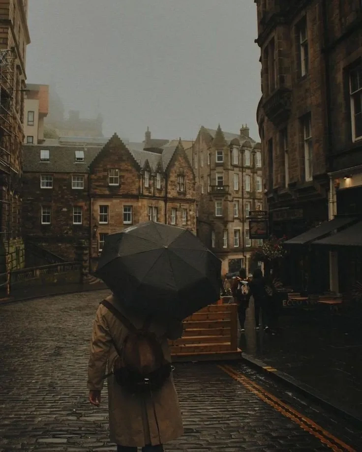 A person viewed from behind walks down a wet, cobblestoned street holding a large black umbrella, wearing a beige trench…
