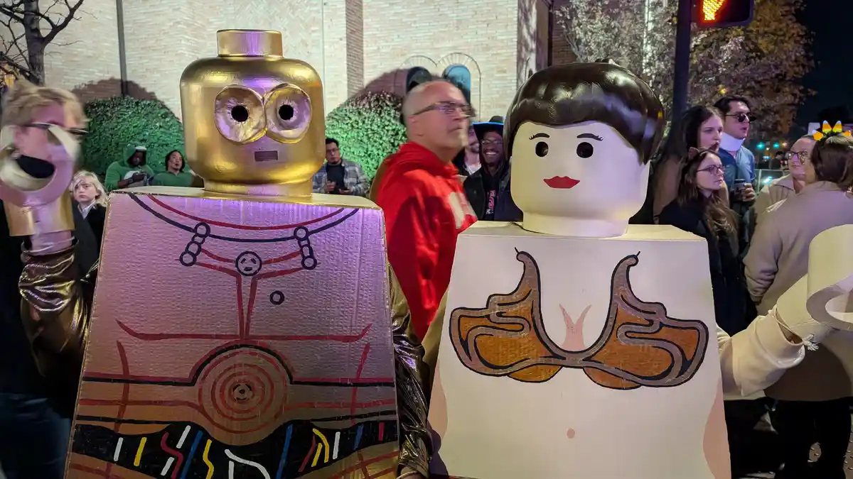 Two people in large, homemade costumes resembling Lego minifigures stand side by side at a nighttime outdoor event. The…