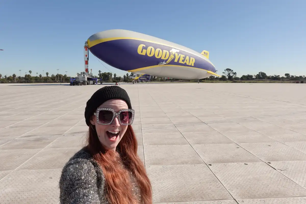 A woman with long red hair, wearing a black beanie and large, sparkly sunglasses, smiles widely with her mouth open in…