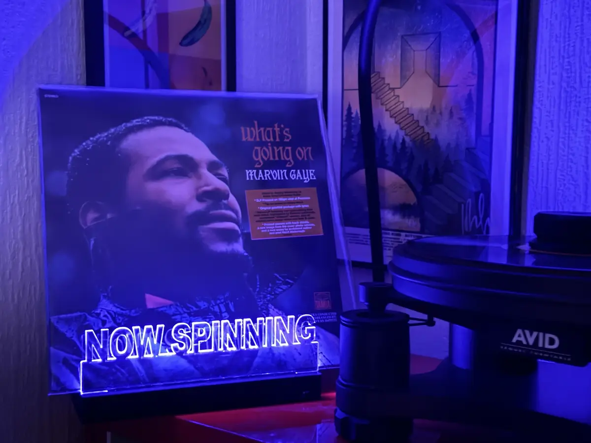 A vinyl record album cover of Marvin Gaye's 'What's Going On' is displayed next to a turntable under blue lighting. The…