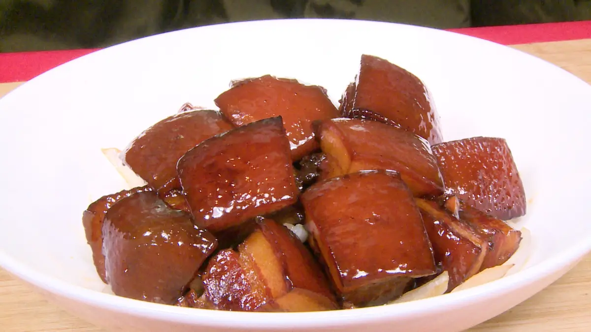 A white bowl filled with glossy, brown, cubed braised pork belly sits on a wooden surface with a red placemat. The overlaid…