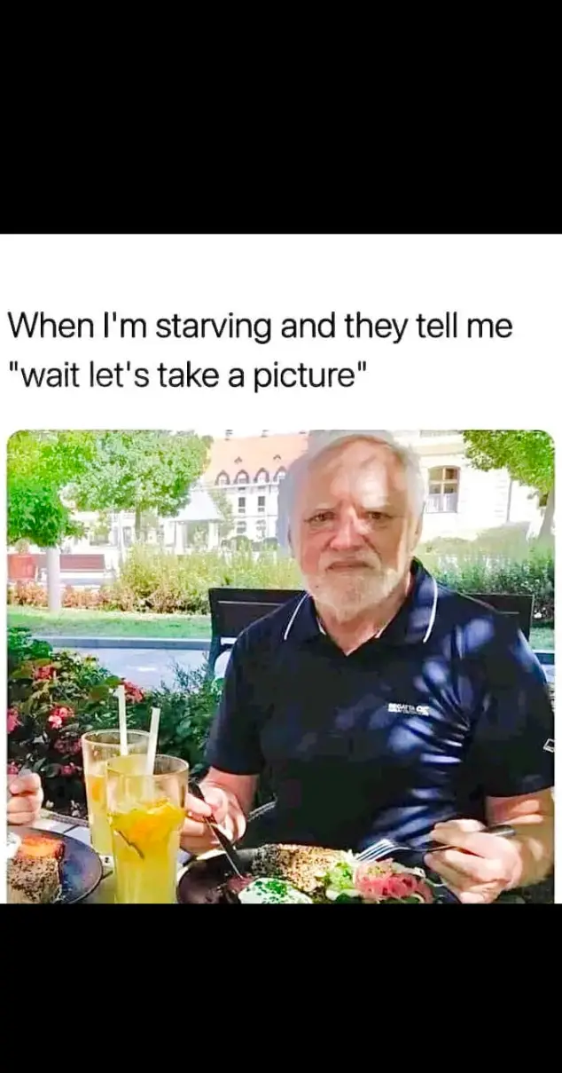 A man with gray hair and a beard, wearing a dark polo shirt, sits at an outdoor table with food and drinks in front of him…