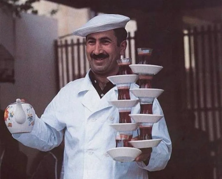 a smiling man with a mustache and white hat wears a white coat while balancing a tall, precarious tower of white saucers and…