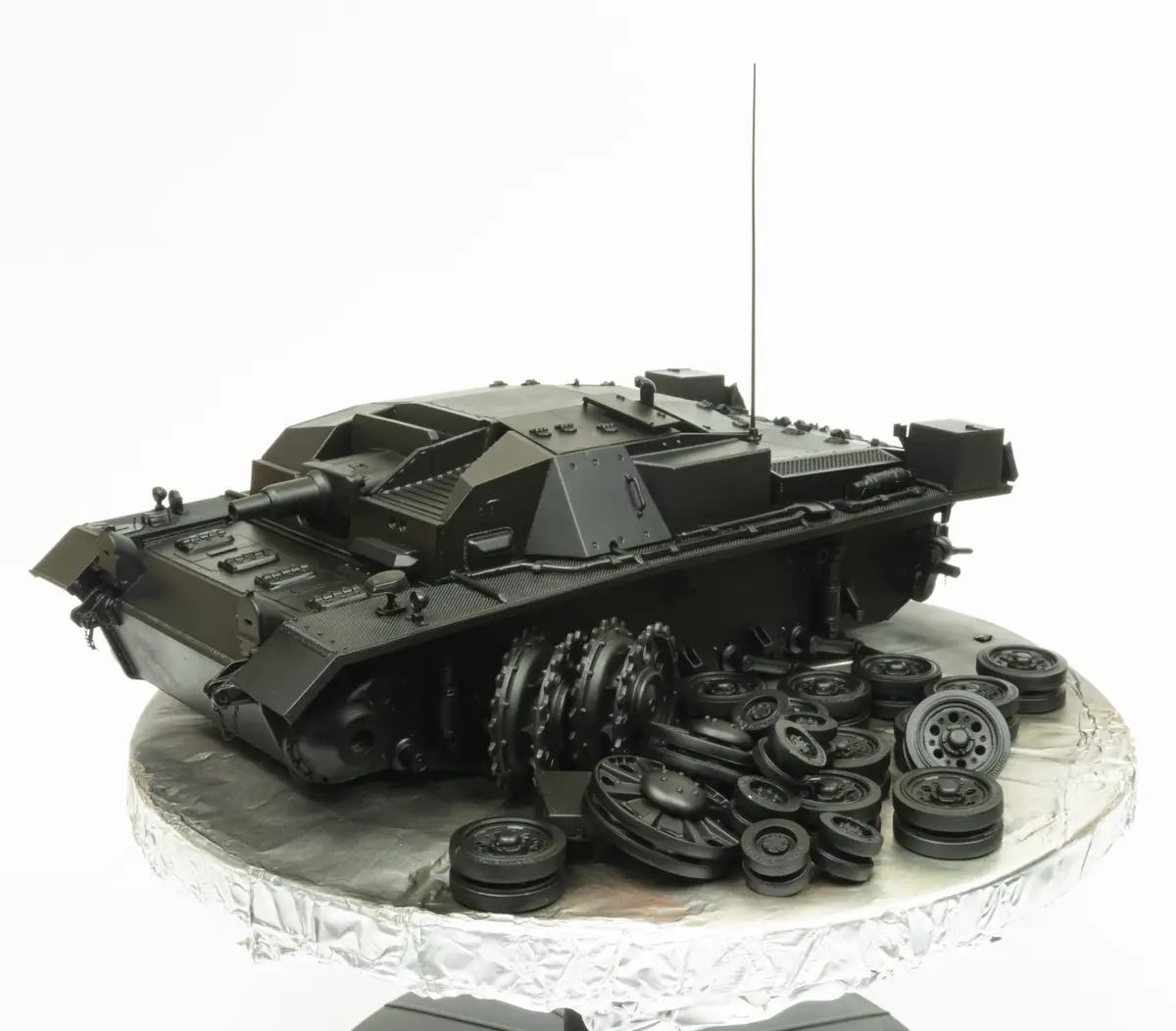 A black cake shaped like a detailed tank model sits on a silver foil-covered cake stand, surrounded by numerous black…