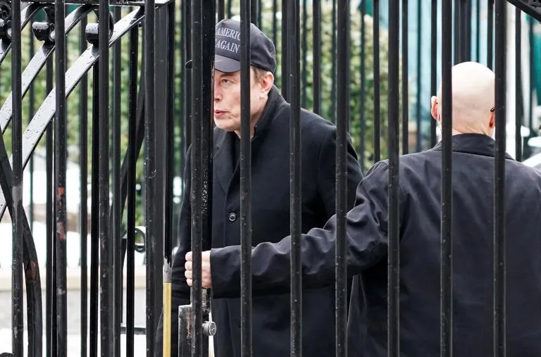 A man wearing a black cap with the text 'AMERICA AGAIN' and a black coat stands behind a black metal gate, looking sideways…