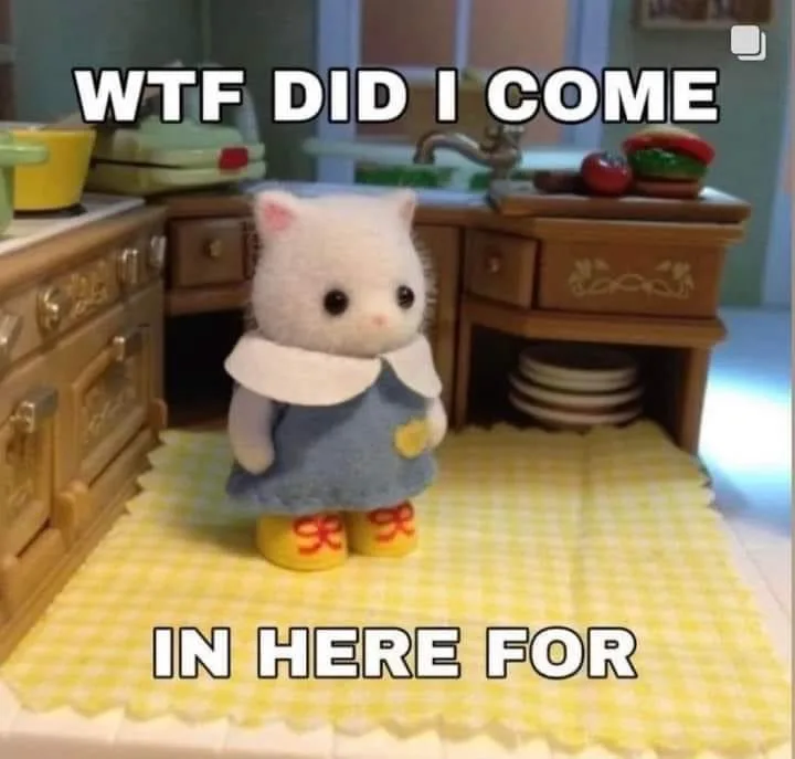 A small, white, plush cat figurine wearing a blue dress and yellow boots stands in a miniature kitchen with wooden cabinets…