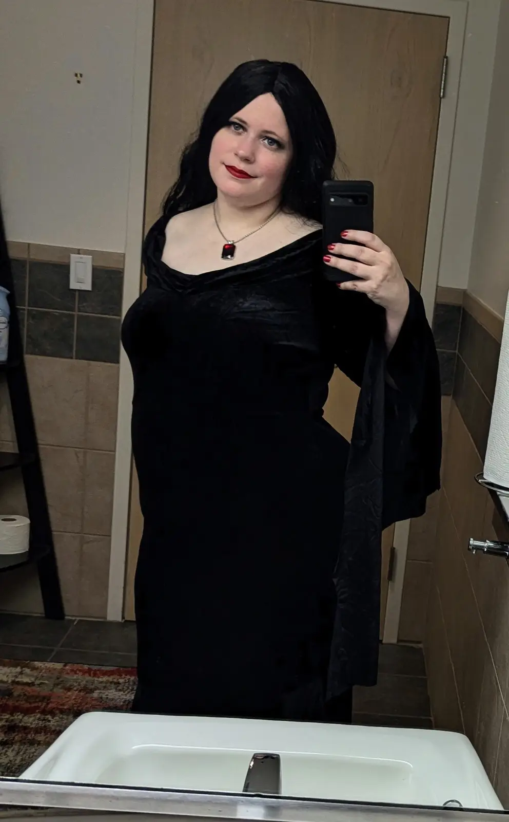 A person with long black hair and red lipstick takes a mirror selfie in a bathroom while wearing a black velvet dress and a…