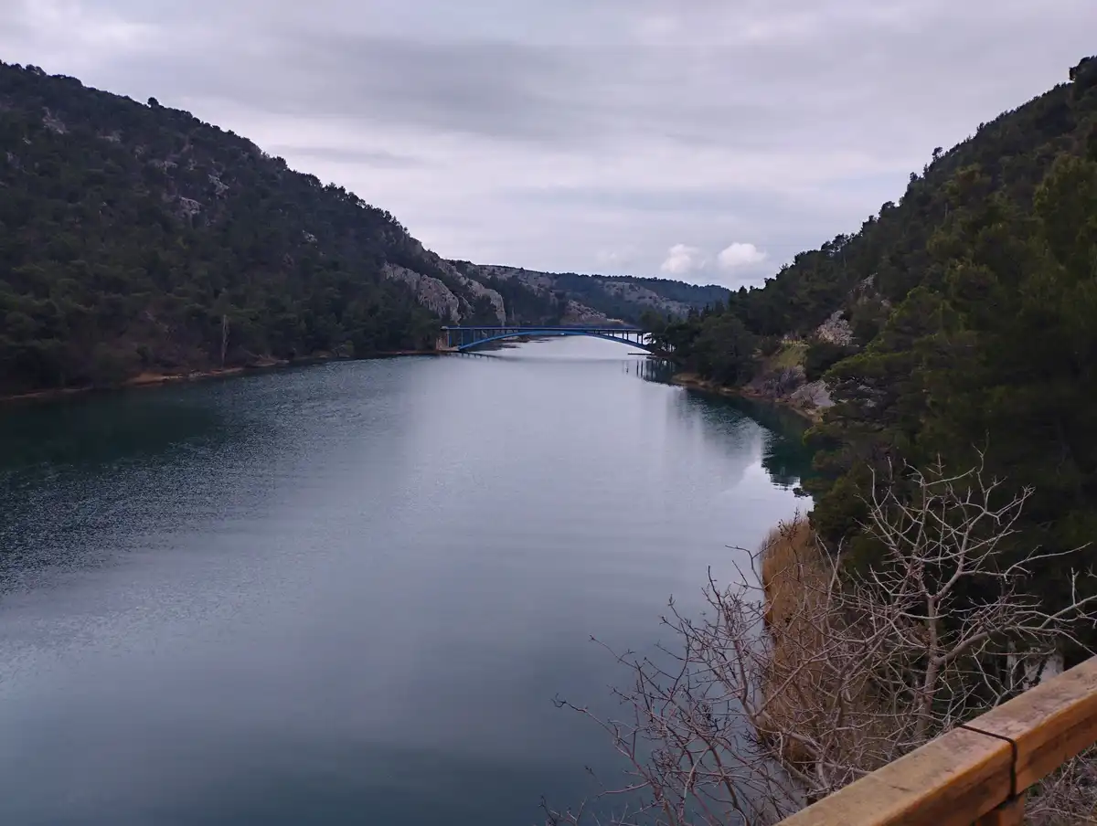 A scenic view of a calm river flanked by forested hills, with a blue arched bridge spanning the water. The photo is taken…