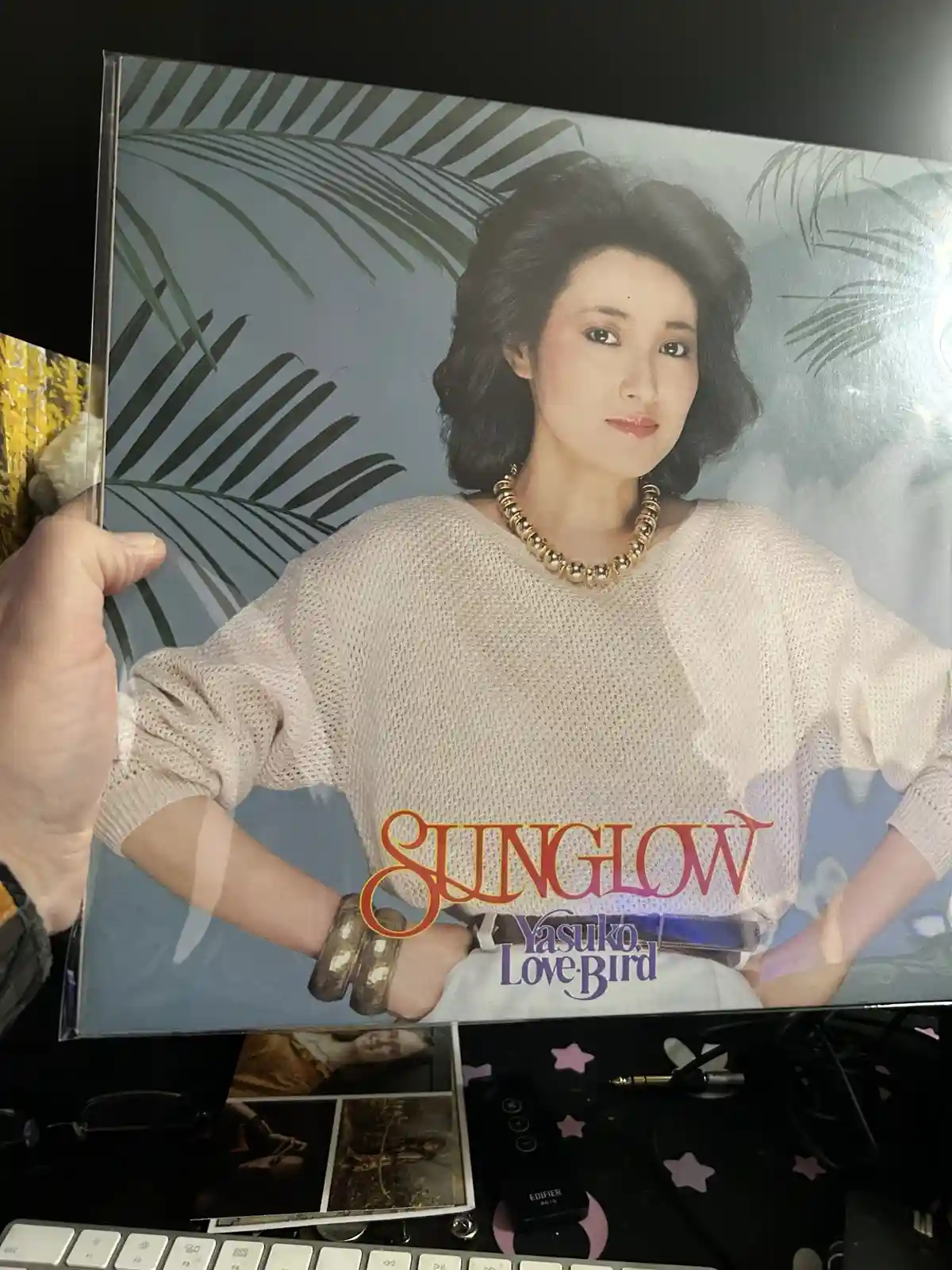 A hand holds up a vintage album cover featuring a woman with dark, styled hair wearing a light-colored knit sweater, a…