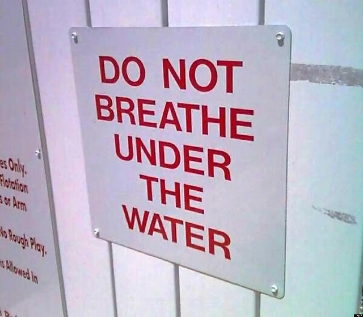 A close-up photo shows a white metal sign with bold red text reading 'DO NOT BREATHE UNDER THE WATER', mounted on a…